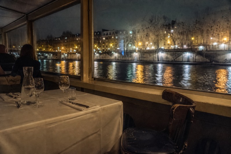 Dinner table set by a window on a Seine river cruise boat with the lit riverbank behind