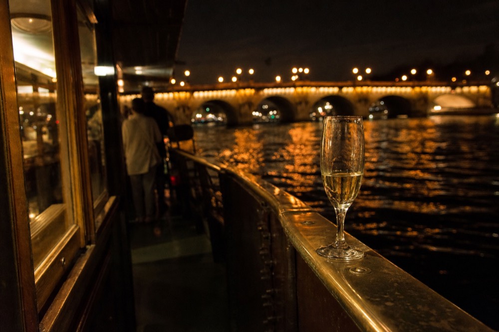 Glass of Champagne on a boat rail with illuminated Paris bridges behind, no text, no logos, no people