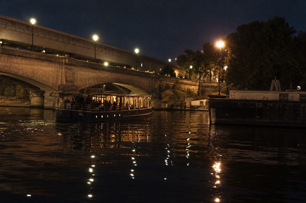 Bistro-style dinner cruise boat gliding past illuminated Parisian bridges at night, no text, no logos, no people