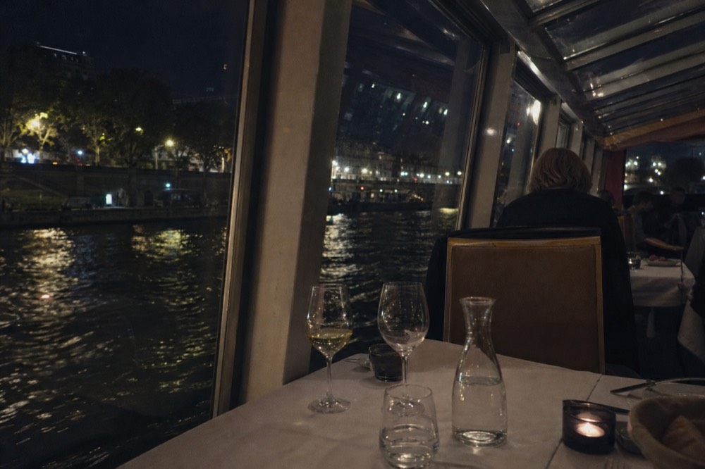Set dinner table with wine glasses inside a glass-enclosed cruise boat on the Seine at night, no text, no logos, no people