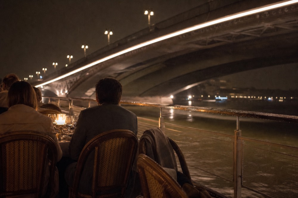 Passengers at a candlelit table on an open terrace of a Seine dinner cruise passing illuminated Parisian bridges, no text, no logos, no people