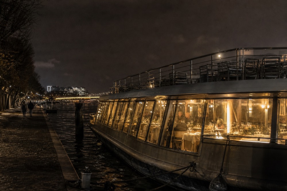 Grand glass-enclosed dinner cruise boat moored on the Seine at night with Paris illuminated behind it, no text, no logos, no people